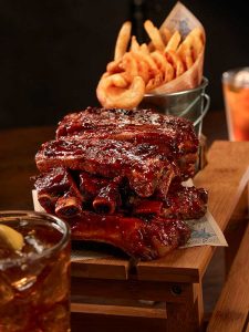 Cafe Hollywood BBQ Ribs smothered with BBQ sauce with hand-cut steak fries on a wooden board with an iced tea